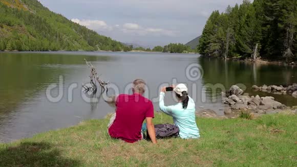 游客夫妇坐在山湖旁用手机拍照视频的预览图
