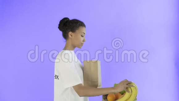 快乐的女人在紫罗兰的背景下装入有机健康蔬菜和水果的纸袋视频的预览图