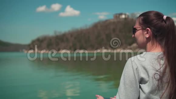 春天的一天一位戴太阳镜的年轻女子坐在湖边在电话上拍照在大自然中放松视频的预览图