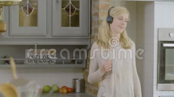 幸福的女人拿着咖啡杯在厨房里听着耳机里的音乐视频的预览图