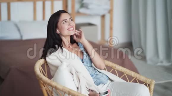 快乐的女人带着格子呢坐在扶手椅上听着耳机上的音乐跳舞视频的预览图