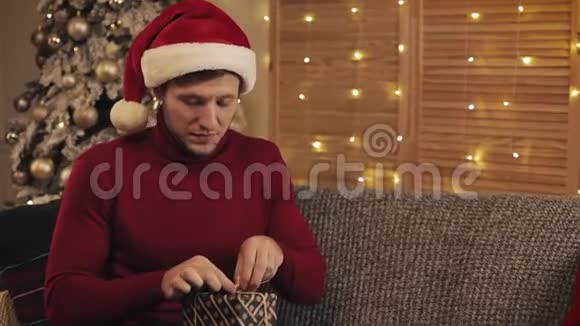 快乐的人戴着圣诞帽和冬衣坐在客厅圣诞树附近的沙发上系着蝴蝶结视频的预览图