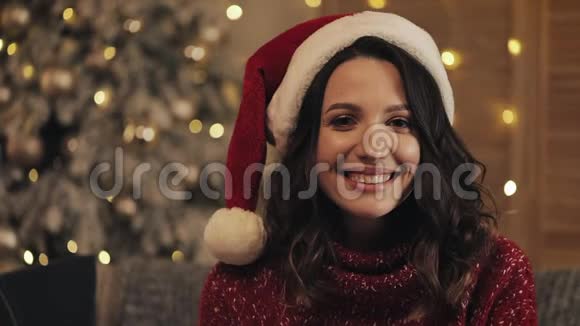 年轻迷人的黑发女人戴着桑塔帽坐在圣诞树上的肖像背景是闪闪发光的灯光视频的预览图