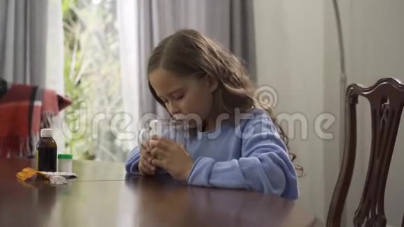 可爱的小女孩一边喝凉茶一边坐在家里的桌子上在厨房里喝茶治感冒的孩子视频的预览图