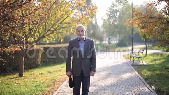 英俊的老人穿上灰色夹克和黑色帽子白发苍苍的胡须老头走在秋天的公园里黄色视频的预览图