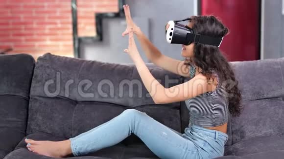 在沙发上戴着虚拟现实模拟眼镜玩游戏的赤脚微笑年轻女子视频的预览图