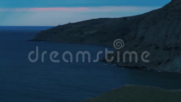 鸟瞰长长的灰色悬崖上面覆盖着灌木和靠近大海的灯塔蓝色和粉红色的夜空视频的预览图