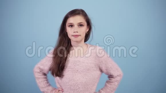 在淡蓝色的摄影棚里摆姿势的小模特棕色头发的女孩看着相机微笑着打手势视频的预览图