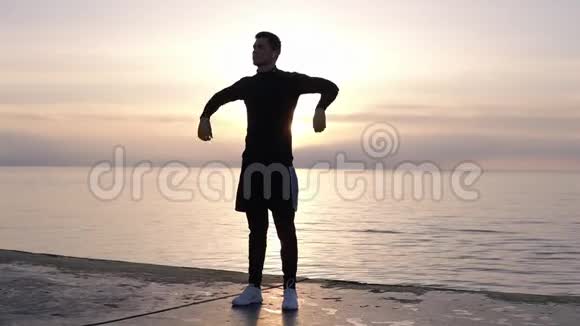 穿短裤的年轻高个子的剪影在靠近大海的木地板上热身伸展着他的双臂视频的预览图