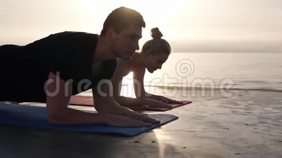 集中的白种人男人和金发女孩在户外训练时在靠近大海的地方在瑜伽垫上拿着木板的位置视频的预览图
