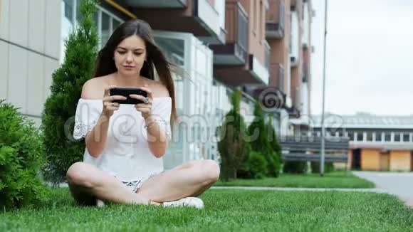 快乐美女在智能手机上玩手机游戏坐在房子附近的草地上视频的预览图