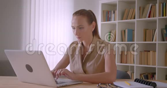 优雅的白种人女商人在笔记本电脑上打字时忙碌地转向相机在办公室里微笑视频的预览图