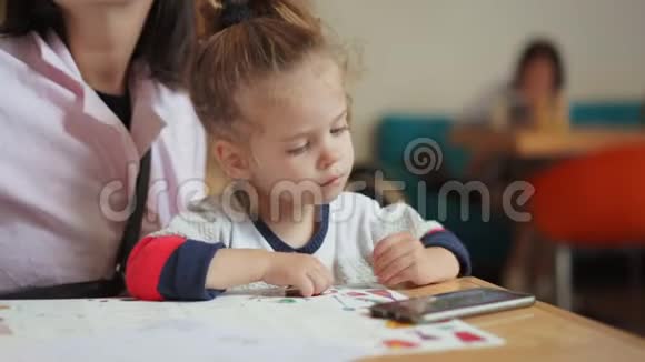 一个欧洲貌美的小女孩正坐在妈妈怀里的一张桌子上看着智能手机上的视频视频的预览图