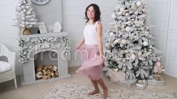美丽的女孩子在圣诞树附近微笑着穿着漂亮的裙子在装饰的房子里跳舞新年快乐视频的预览图