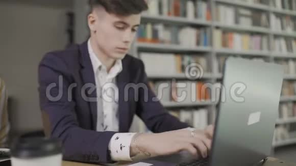 年轻英俊的时髦男学生正坐在轻巧的大图书馆里面前放着笔记本电脑视频的预览图