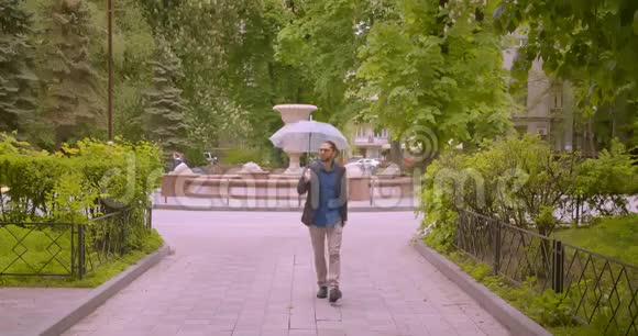 英俊的时髦商人带着雨伞微笑着走进公园里的摄像机正对着镜头视频的预览图