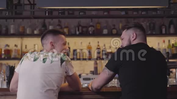 两个人坐在酒吧柜台的后景第三个加入公司的家伙朋友拥抱放松的男性朋友视频的预览图