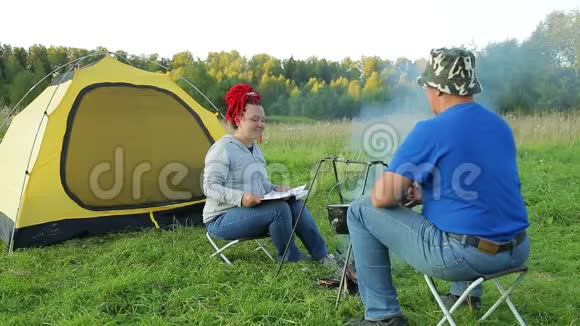 一个男人和一个女人在日落时靠近一个靠近火的帐篷在锅里做晚餐视频的预览图