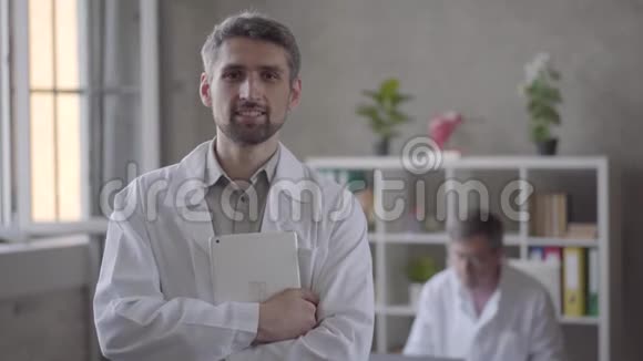 一幅英俊微笑的医生拿着平板电脑在镜头中看着他的同事的照片视频的预览图