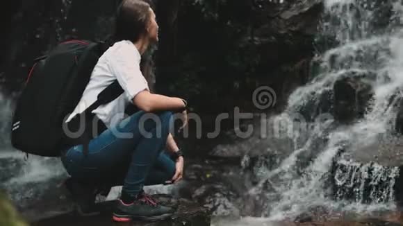 女徒步旅行者蹲伏在狂野的飞溅瀑布附近视频的预览图