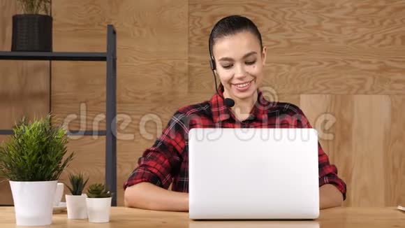 美丽的自由职业者女性摆姿势和工作与耳机与麦克风在一个小办公室桌面或家庭视频的预览图