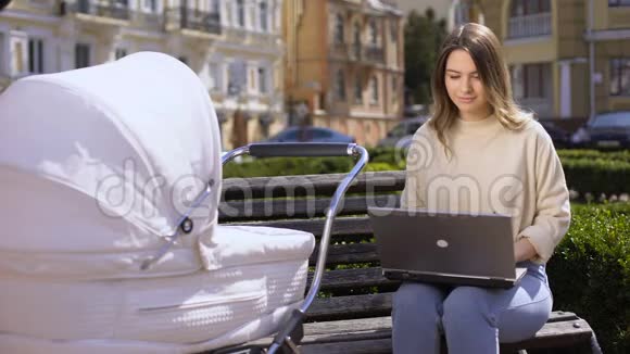 快乐的现代妈妈在手提电脑上看着婴儿坐在马车上的长椅上视频的预览图
