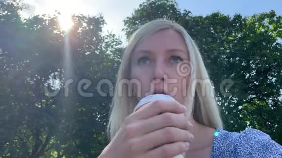 年轻的金发白种人女人坐在夏季城市公园的长椅上用一次性吸管喝咖啡视频的预览图