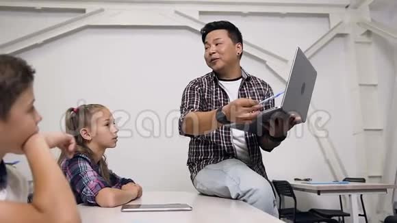 年轻的韩国老师坐在桌子上手里拿着笔记本电脑给学生们上课小学学生视频的预览图