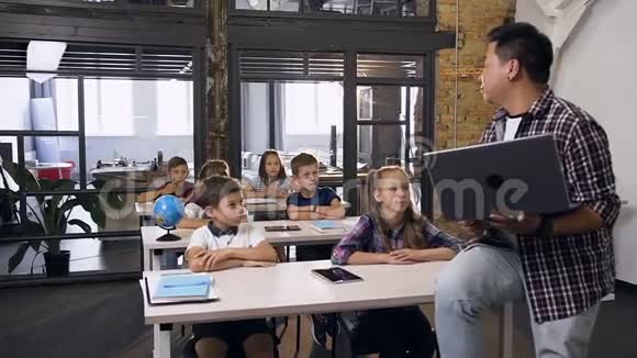 年轻的韩国男教师坐在桌子上手提电脑为六名小学生上课学龄儿童视频的预览图