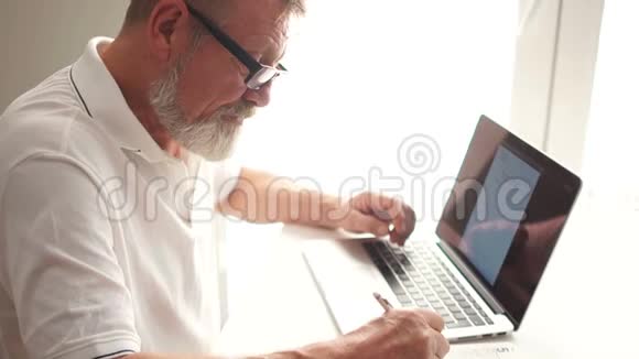 这位蓄着胡子的作家正借助笔记本电脑写一本书成熟的男人在家里用笔记本电脑工作视频的预览图