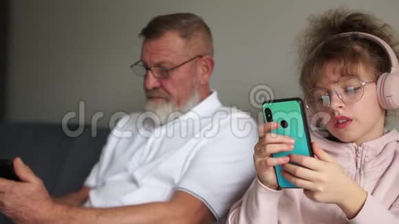 一个十几岁孩子的祖父和孙女坐在沙发旁边每个人都在看他们的智能手机视频的预览图