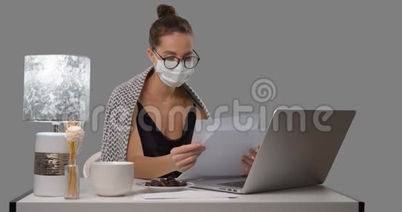 戴着医用口罩的女病人在家工作她坐在桌子旁阅读纸质文件在笔记本电脑上打字视频的预览图