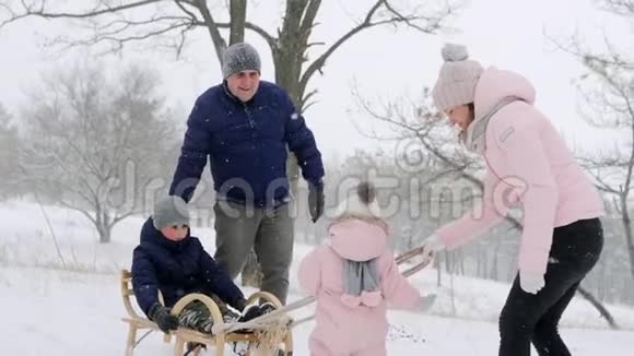 幸福的家庭在下雪的冬天滑雪女儿帮助父亲和母亲在下雪时和儿子拉雪橇男孩视频的预览图