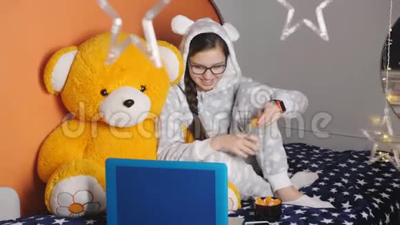 女孩少年穿着睡衣坐在床上抱着一只橙色的大泰迪熊在我的笔记本电脑上看电影视频的预览图