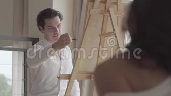 英俊的男艺术家在家画漂亮的半着装女人年轻漂亮的模特为画的男人摆姿势视频的预览图