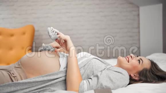 快乐美丽的年轻孕妇玩着小可爱的婴儿鞋躺在床上视频的预览图