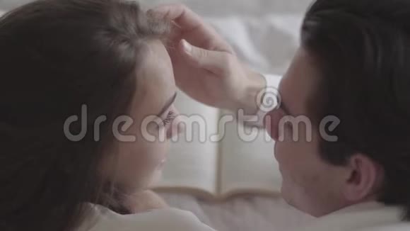 一对幸福的年轻夫妇躺在床上看书特写英俊的男人摸着女人的脸投标视频的预览图