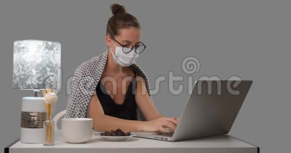 生病的女人在家里自由工作她坐在桌子上用一张毯子盖着医疗面罩隔离在灰色的BG上视频的预览图
