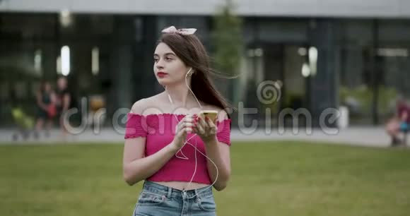 美丽的年轻女子用智能手机打字听音乐在公园里散步视频的预览图