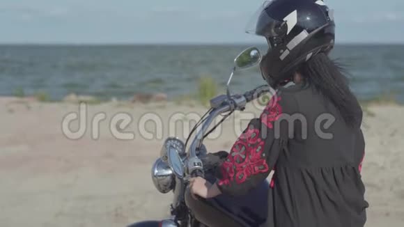 白人女孩穿着黑色连衣裙和头盔坐在摩托车上看着相机爱好旅游和视频的预览图
