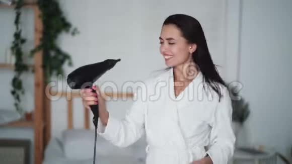 年轻女子穿着白色浴袍跳舞用吹风机吹干头发视频的预览图