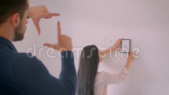 白种人黑发女孩在白墙上选择相框照片的位置而她的男朋友则用相框造型视频的预览图