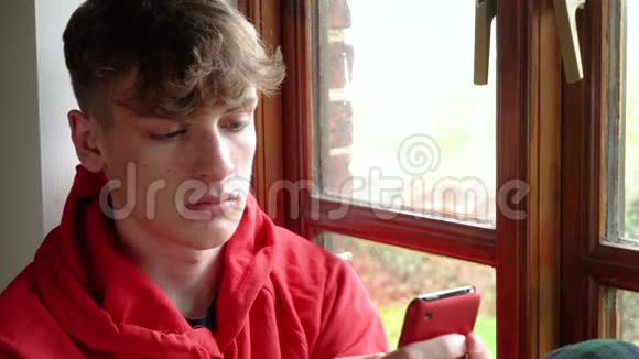 伤心沮丧的青少年男性青少年年轻人望着窗外用手机社交媒体视频的预览图