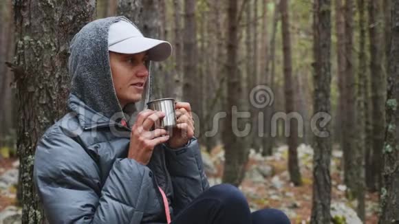 年轻女子徒步旅行者坐在森林里喝金属杯里的茶的肖像视频的预览图