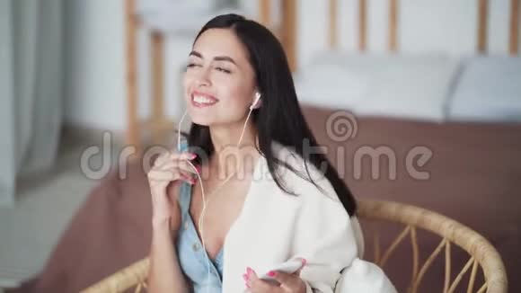 快乐的女人带着格子呢坐在扶手椅上听着耳机上的音乐跳舞视频的预览图