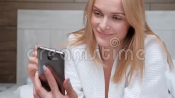 一个年轻迷人的中年金发女人穿着白色浴袍躺在酒店房间的床上在视频的预览图