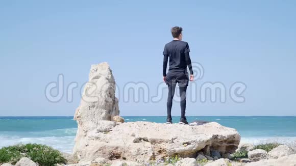 年轻健康的白种人站在海滩上的大石头上戴上太阳镜开始跑步慢动作视频的预览图