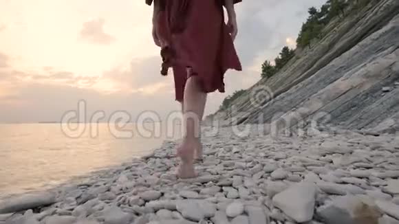 一个年轻女孩的腿特写她穿着一件红色的裙子从后面走在日落时海边的岩石海滩上A视频的预览图