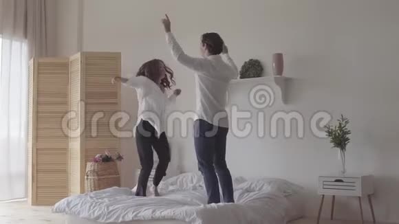 快乐的一对夫妇在家里或酒店的房间里一起在床上跳舞年轻女子高兴地跳来跳去视频的预览图