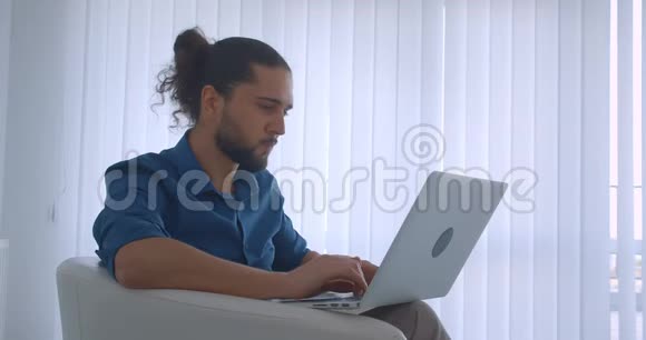 现代自由职业者在光办公室坐在扶手椅上的笔记本电脑上打字的照片变成了相机和微笑视频的预览图